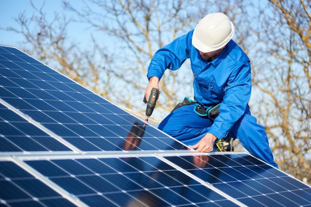 Male engineer in blue suit and protective helmet installing photovoltaic panel system using screwdriver. Professional electrician mounting solar module on roof. Alternative energy ecological concept.